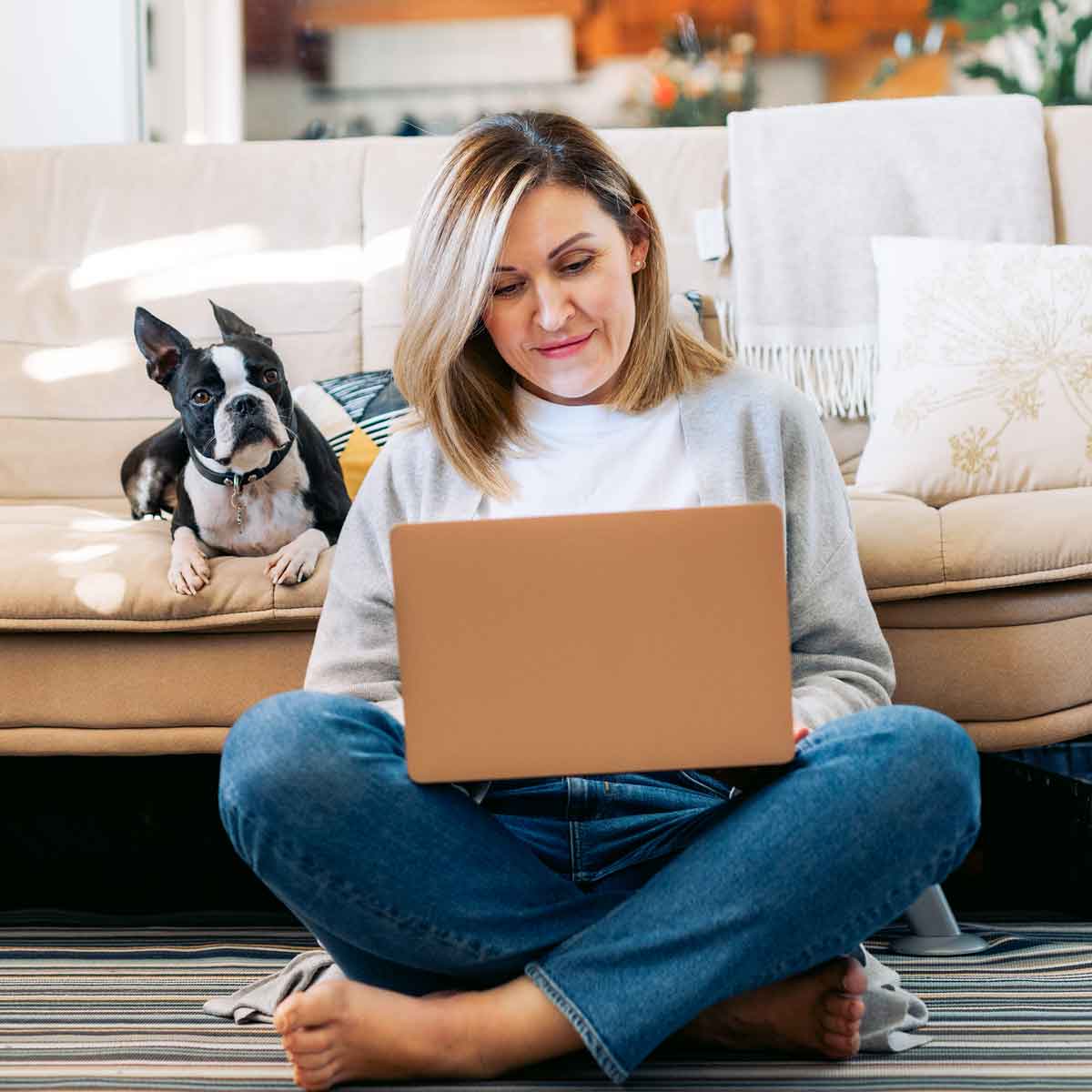 Women sitting on the floor with her dog on the couch behind her looking at her laptop