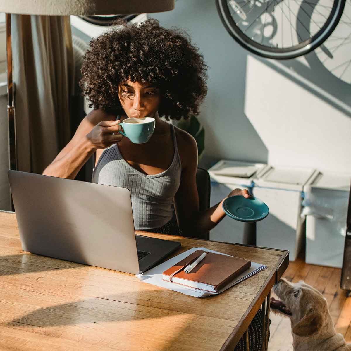 African American woman at a desk looking at her computer drinking a cup of coffee with her dog looking up at her.